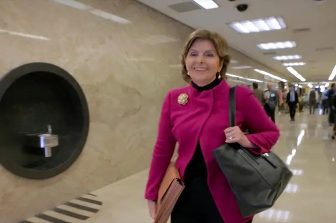 Gloria Allred, in a pink suit jacket, smiles as she walks down a court building hallway